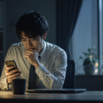 夜のオフィスでスマホを見ながら退職代行を使うべきか悩む会社員