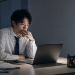 夜のオフィスでノートパソコンを見つめながら、向いていない仕事を辞めるべきか悩む会社員