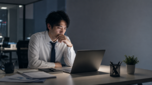 夜のオフィスでノートパソコンを見つめながら、向いていない仕事を辞めるべきか悩む会社員
