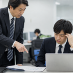 オフィスで上司に厳しく指摘され、困った表情でパソコンを見る若手社員
