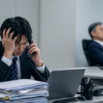 トラブル対応の電話を一人で抱え、困った表情を見せる会社員と、後方で対応しない上司